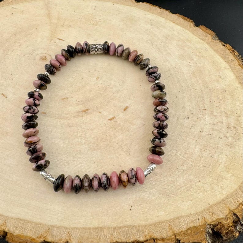 May include: A beaded bracelet featuring pink and black gemstone beads. The bracelet has a silver clasp and a decorative silver bead in the center. The beads are oval-shaped and strung together to form a complete circle.