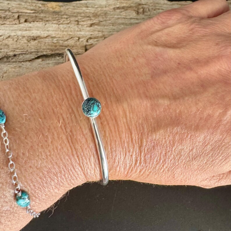 May include: A silver-toned cuff bracelet with a single, round turquoise stone. The bracelet is simple and elegant, with a smooth, polished finish. The turquoise stone is a vibrant blue-green color, adding a pop of color to the design.