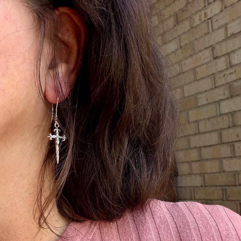 May include: A silver-toned dangle earring featuring a small, ornate cross design with intricate detailing. The earring hangs from a thin metal hook and has a pointed, dagger-like extension at the bottom, creating a slender and elongated silhouette.