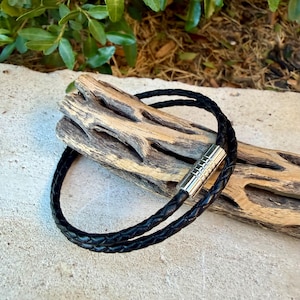 May include: A black braided leather bracelet featuring a cylindrical silver-toned metal magnetic clasp. The bracelet is wrapped around a piece of light-colored, weathered driftwood with a textured, porous surface and visible natural grooves.