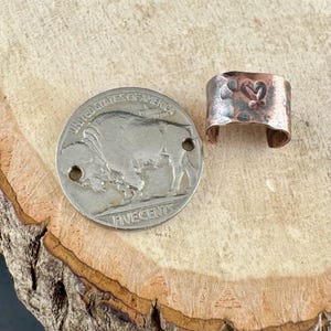 May include: A silver nickel coin with a buffalo image next to a hammered copper ring with a heart design. The ring is open at the bottom and has a rustic, textured appearance.