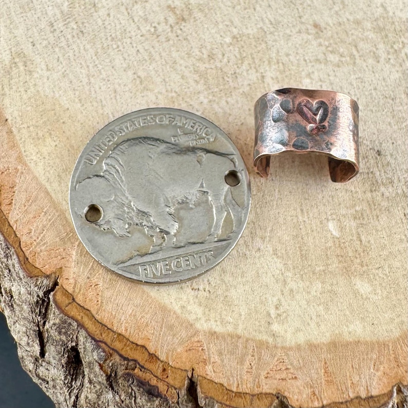 May include: A silver nickel coin with a buffalo image next to a hammered copper ring with a heart design. The ring is open at the bottom and has a rustic, textured appearance.