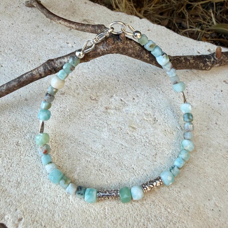 Puede incluir: Una pulsera de cuentas con peque&ntilde;as piedras preciosas facetadas en tonos azul claro y verde, intercaladas con dos cuentas espaciadoras de metal plateado ornamentado. La pulsera incluye un cierre de mosquet&oacute;n y una anilla de metal plateado.
