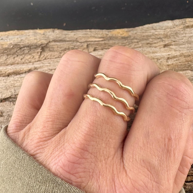 May include: Three gold-colored wave rings are stacked on a finger. The rings have a delicate, undulating design, creating a flowing, organic look. The rings are displayed against a natural wood background.