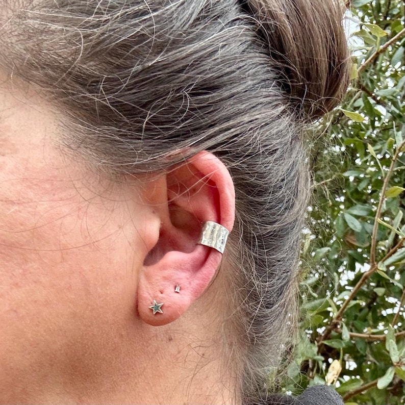 May include: A close-up of an ear adorned with a silver ear cuff and two small stud earrings. The ear cuff has a textured surface. One earring is a star shape, and the other is a small square.
