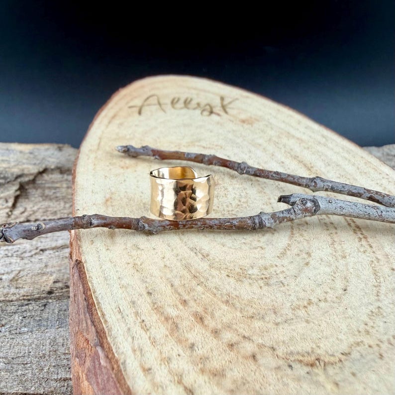 Puede incluir: Un anillo dorado con textura martillada. El anillo ancho se encuentra sobre una superficie de madera con veta natural. Dos peque&ntilde;as ramas se colocan cerca del anillo.