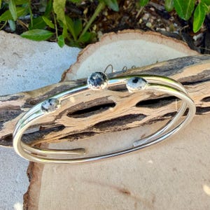 May include: A silver-toned cuff bracelet with three round, speckled stones. The bracelet has a double-band design and is displayed on a piece of driftwood. The stones have a black and white pattern.