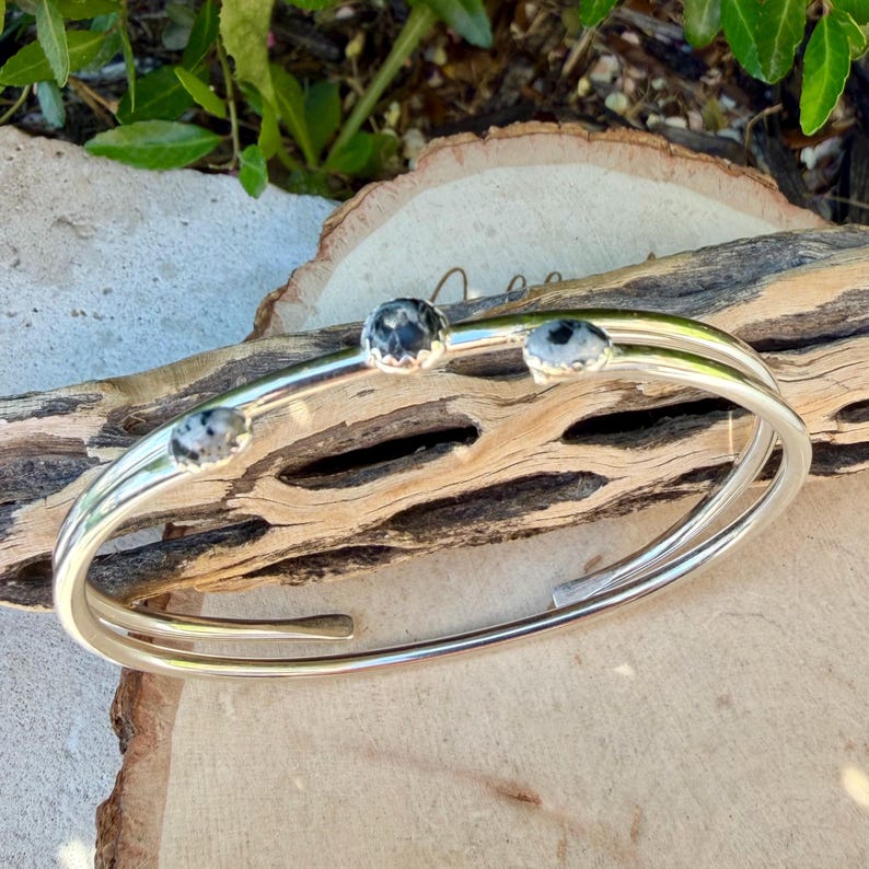 May include: A silver-toned cuff bracelet with three round, speckled stones. The bracelet has a double-band design and is displayed on a piece of driftwood. The stones have a black and white pattern.