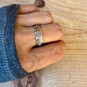 Puede incluir: Un anillo de metal plateado que presenta una banda central con un patr&oacute;n floral grabado y bordes lisos y elevados. El anillo tiene un acabado pulido y un ancho constante, mostrando detalles bot&aacute;nicos intrincados grabados en la superficie.