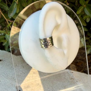 May include: A silver-toned ear cuff featuring a hammered texture and a central embossed sun motif with radiating rays. The metal band is wide and wraps around the outer edge of a white, ear-shaped display stand, showcasing a metallic finish.