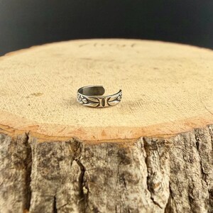 K&ouml;nnte beinhalten: Ein silberfarbener Metallring mit offenem Banddesign. Die Oberfl&auml;che des Rings ist mit einem gepr&auml;gten Muster aus kleinen Blumen und Bl&auml;ttern verziert, was f&uuml;r ein strukturiertes Aussehen rund um den gesamten Ringumfang sorgt.