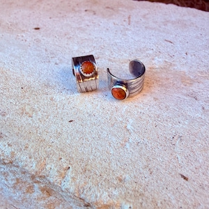 May include: Two silver cuff rings featuring a textured, grooved band design. Each ring is set with a single, round, orange-colored stone in a bezel setting. The metal has a polished finish with visible vertical lines etched into the surface.