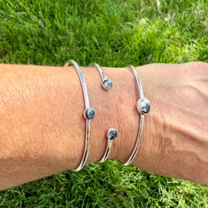 May include: Four silver-toned, open cuff bracelets. Each bracelet features a small, round, black and white stone. The bracelets are displayed on a person's arm against a background of green grass.