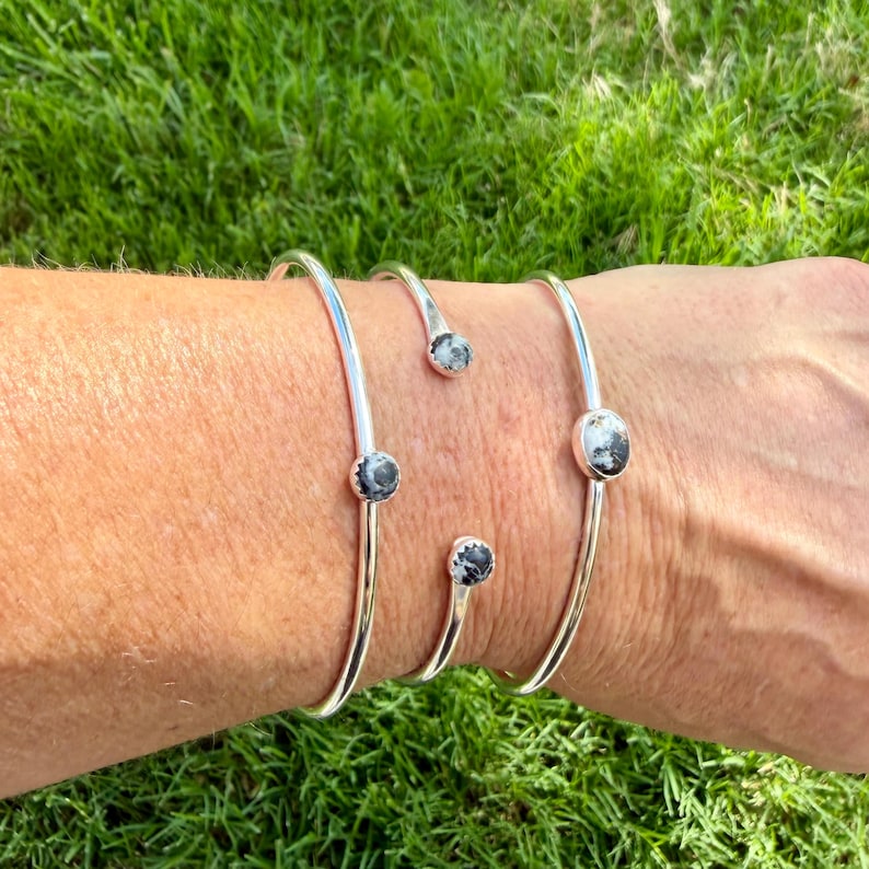 May include: Four silver-toned, open cuff bracelets. Each bracelet features a small, round, black and white stone. The bracelets are displayed on a person's arm against a background of green grass.