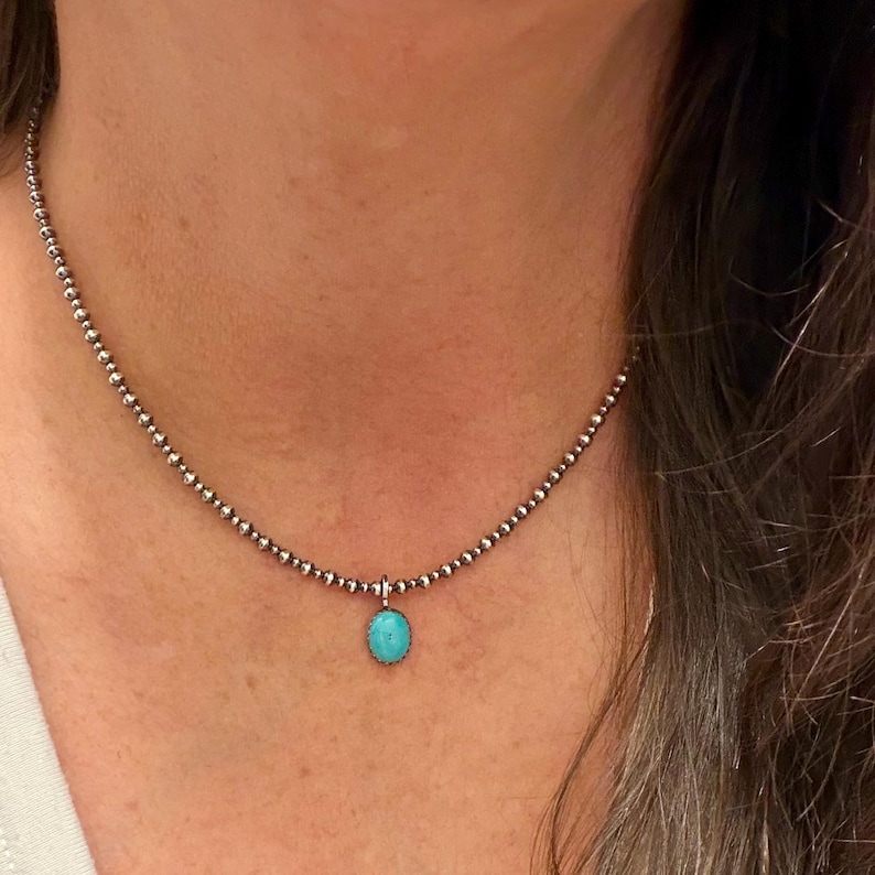 May include: A delicate silver necklace featuring a beaded chain and a small, oval turquoise pendant. The necklace is worn around the neck, showcasing the vibrant blue-green stone. The chain is made of small, round beads.