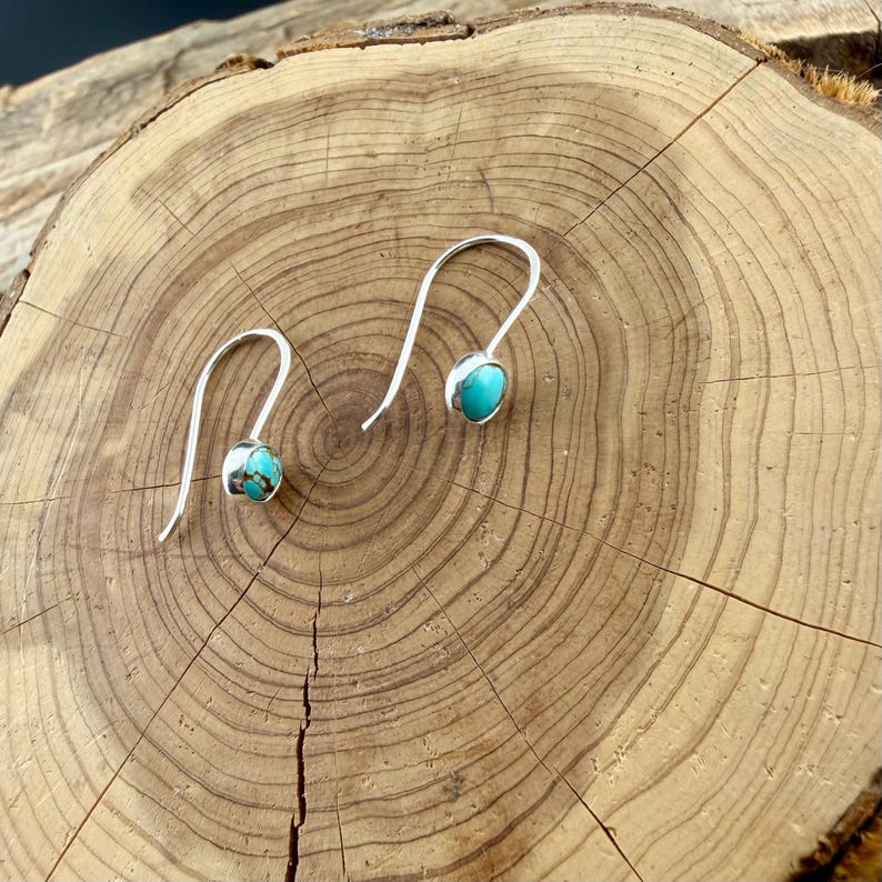 May include: A pair of silver hook earrings, each featuring a small, round, polished turquoise stone with brown matrix veining. The earrings have a simple, curved wire design that allows them to hang freely, showcasing the bright blue-green color.