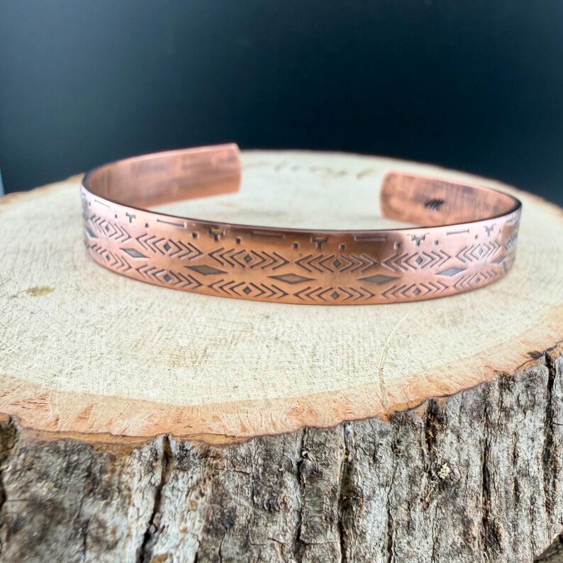 May include: A copper cuff bracelet featuring a repeating geometric pattern of etched diamonds, chevrons, and horizontal lines. The metal has a warm, metallic finish with dark, recessed detailing within the engraved markings along the band.