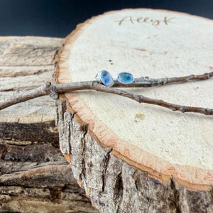 May include: A pair of oval-shaped stud earrings with a silver setting. One earring features a blue stone, and the other has a blue-green stone. The earrings rest on a small branch, set against a wooden background.