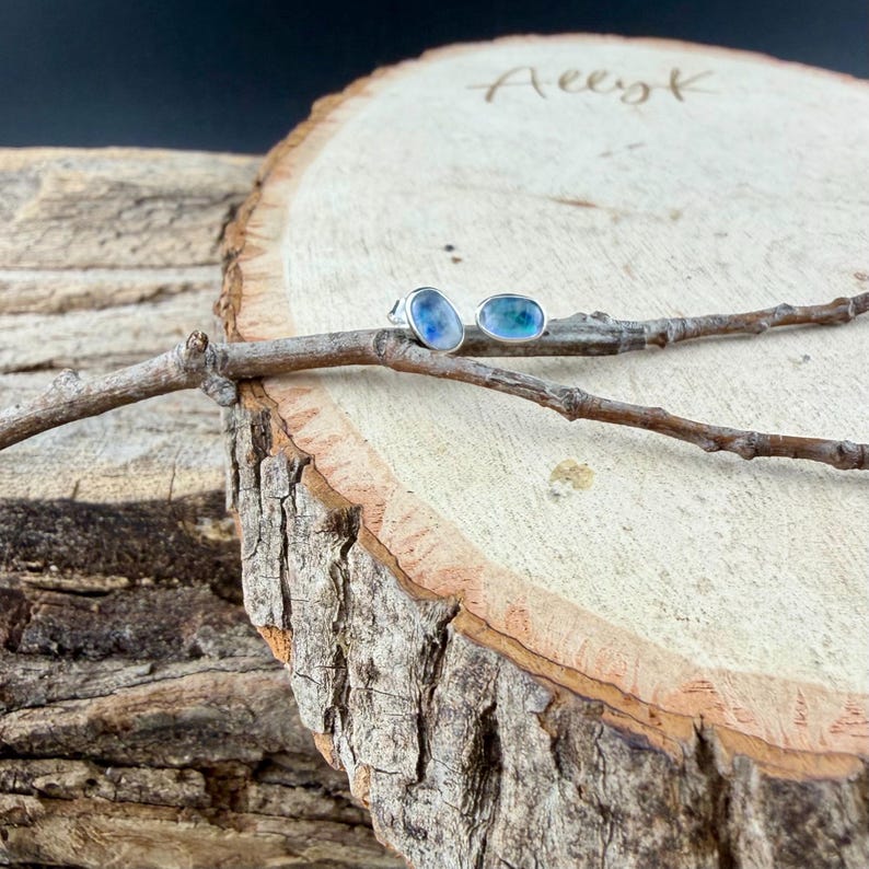 May include: A pair of oval-shaped stud earrings with a silver setting. One earring features a blue stone, and the other has a blue-green stone. The earrings rest on a small branch, set against a wooden background.