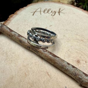 May include: A stack of three silver rings. The top ring has a series of small, round cutouts. The middle ring features a row of small skull designs. The bottom ring has etched text. The rings are on a wooden surface with the name "Allyk" written on it.
