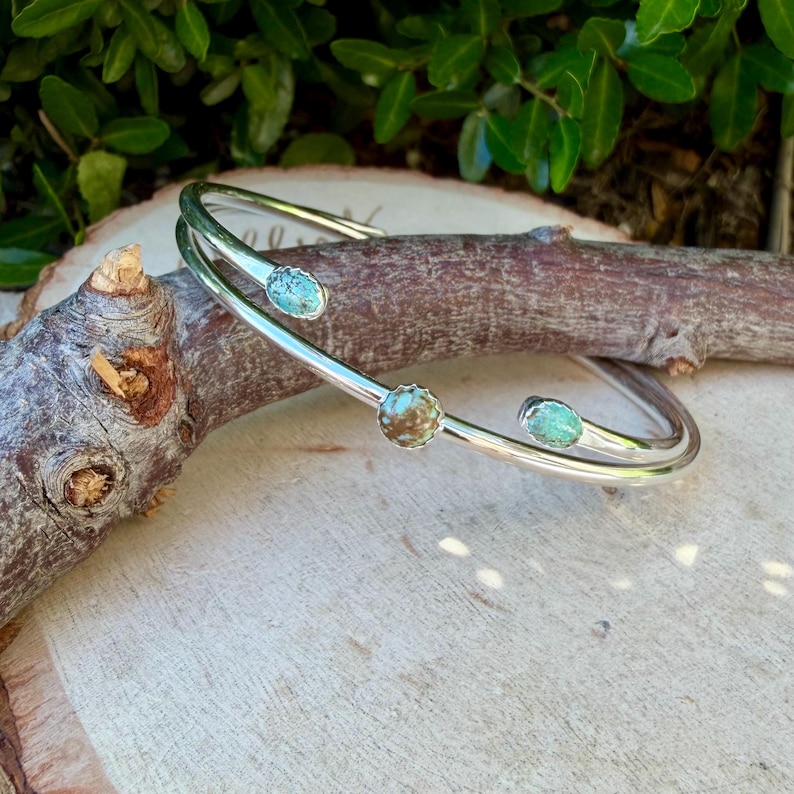 May include: A silver-toned cuff bracelet with three turquoise-colored stones. The bracelet has a double-band design and is displayed on a wooden surface with green foliage in the background. The stones are oval-shaped and set in the metal.