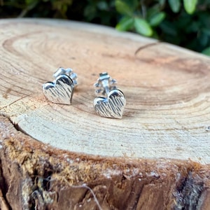 May include: A pair of silver heart-shaped stud earrings featuring a textured, brushed finish on the front surface. Each earring includes a standard post and a secure butterfly clutch backing, showcasing a metallic sheen and detailed lines.