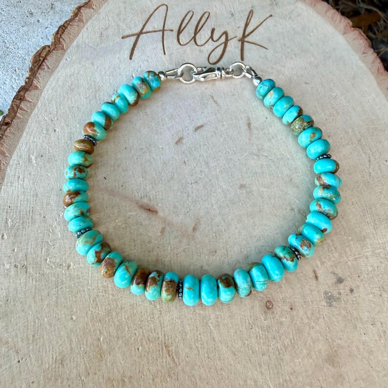 May include: A beaded bracelet featuring round, turquoise-colored stones with brown matrix patterns, accented by small silver-toned metal spacers and a silver-toned lobster claw clasp. The word Ally K is written in cursive script above the bracelet.