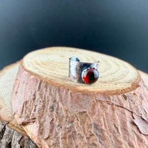 Puede incluir: Un anillo plateado con textura martillada y una gema roja grande y redonda. El anillo se presenta sobre una rodaja de madera natural, creando una est&eacute;tica r&uacute;stica y elegante. La gema es de un color rojo intenso y rico.