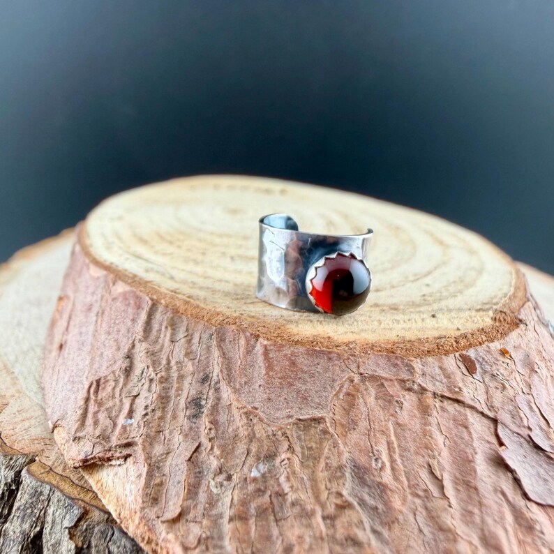 Puede incluir: Un anillo plateado con textura martillada y una gema roja grande y redonda. El anillo se presenta sobre una rodaja de madera natural, creando una est&eacute;tica r&uacute;stica y elegante. La gema es de un color rojo intenso y rico.