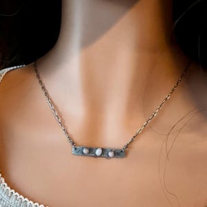 May include: A silver-toned necklace featuring a rectangular bar pendant with a textured, dark finish. The bar is set with three small, oval-shaped, iridescent white stones arranged horizontally along the center of the metal piece.