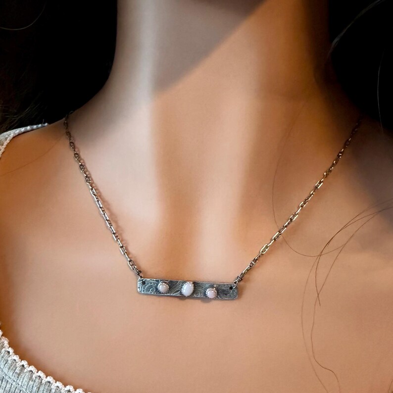 May include: A silver-toned necklace featuring a rectangular bar pendant with a textured, dark finish. The bar is set with three small, oval-shaped, iridescent white stones arranged horizontally along the center of the metal piece.