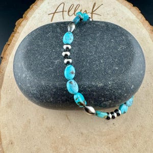 May include: A turquoise beaded bracelet with silver-colored accents. The bracelet features oval-shaped turquoise beads and small, round silver beads. The bracelet is displayed on a dark gray stone, with the word "Alluk" in cursive script.