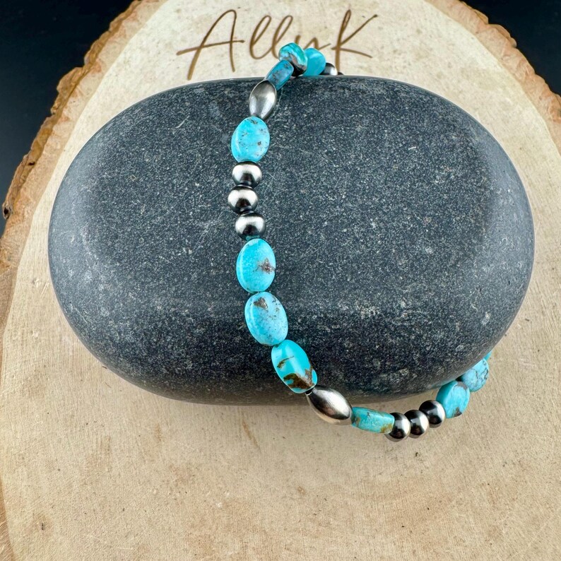 May include: A turquoise beaded bracelet with silver-colored accents. The bracelet features oval-shaped turquoise beads and small, round silver beads. The bracelet is displayed on a dark gray stone, with the word "Alluk" in cursive script.