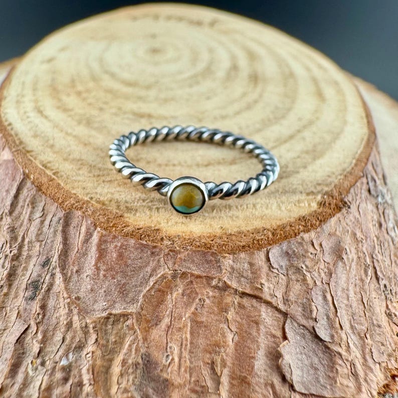 May include: A silver-toned ring with a twisted band design. The ring features a small, round, light brown gemstone at the center. The ring is displayed on a wooden surface.