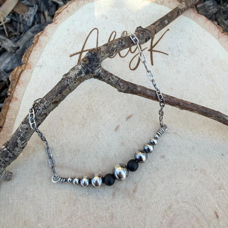 May include: A silver necklace with a chain and a series of metal beads. The beads vary in size, with larger, polished silver spheres and smaller, matte black beads. The necklace is displayed on a wooden surface with a branch.