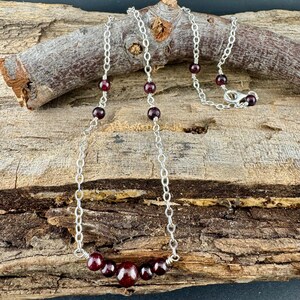 May include: A silver chain necklace with dark red garnet beads. The necklace features a series of small, round garnet beads interspersed along the chain, with a cluster of larger beads at the center. The necklace is displayed on a piece of wood.