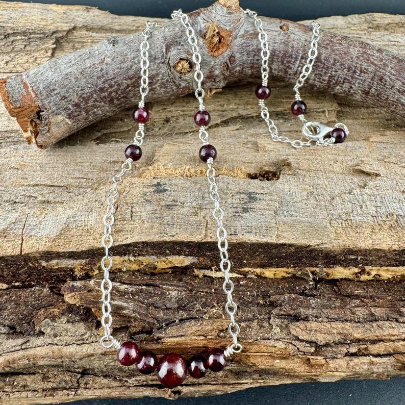 May include: A silver chain necklace with dark red garnet beads. The necklace features a series of small, round garnet beads interspersed along the chain, with a cluster of larger beads at the center. The necklace is displayed on a piece of wood.