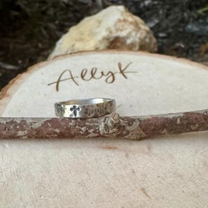 May include: A silver ring with a hammered texture and a cross design. The ring is displayed on a wooden branch, with the name "Allyk" written on a wooden slice in the background. The ring is a piece of jewelry.