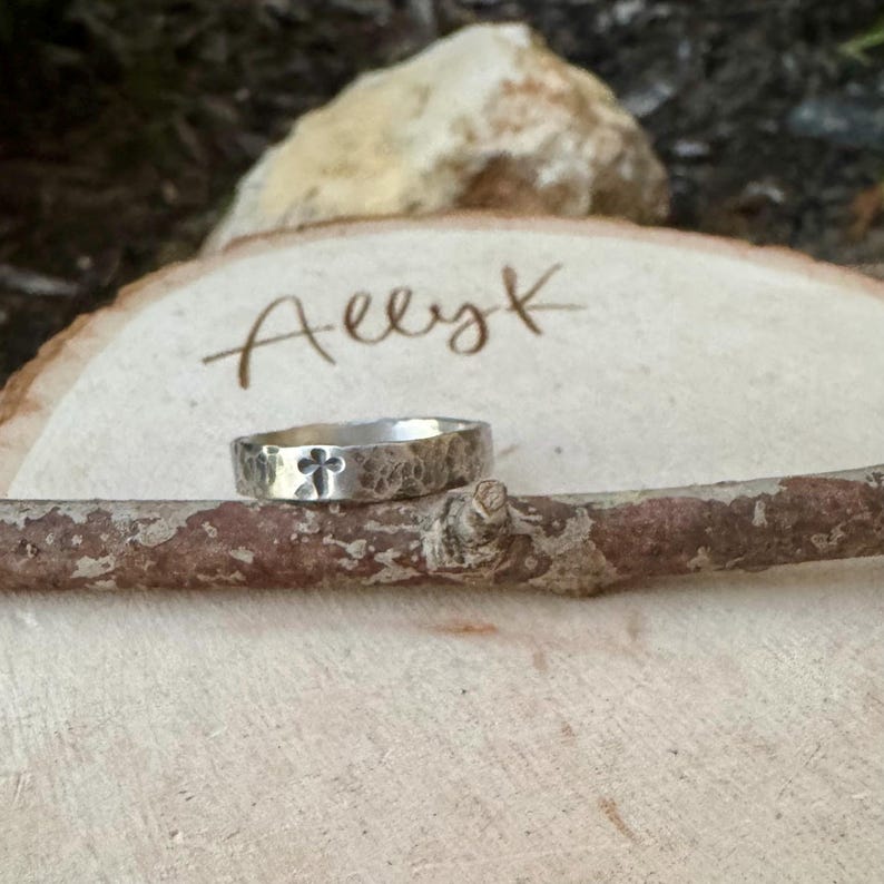 May include: A silver ring with a hammered texture and a cross design. The ring is displayed on a wooden branch, with the name "Allyk" written on a wooden slice in the background. The ring is a piece of jewelry.