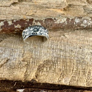 May include: A silver ring with a textured, organic design. The ring's surface has a detailed, irregular pattern, possibly resembling a natural element. The ring is set against a wooden background, highlighting its metallic sheen.