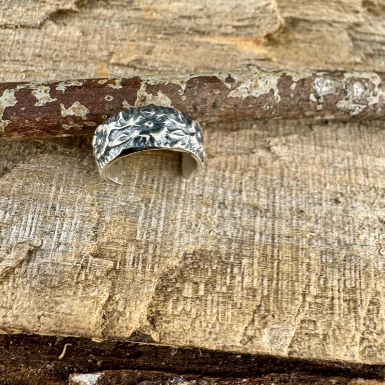 May include: A silver ring with a textured, organic design. The ring's surface has a detailed, irregular pattern, possibly resembling a natural element. The ring is set against a wooden background, highlighting its metallic sheen.