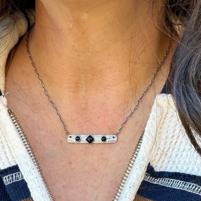 Pu&ograve; includere: Una collana a barra color argento con superficie testurizzata e tre pietre nere. La pietra centrale &egrave; quadrata, affiancata da due pietre nere rotonde pi&ugrave; piccole. Il ciondolo &egrave; sospeso a una sottile catenina color argento.