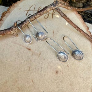 May include: Two pairs of silver-toned drop earrings featuring circular, scalloped-edge charms with a sunburst-style embossed pattern. The earrings have long, curved wire hooks that extend from the center of each decorative metal disc.