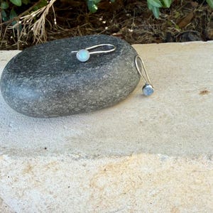 May include: A pair of silver earrings featuring round, iridescent moonstone cabochons set in metal bezels. The earrings have long, curved silver ear wires that extend from the back of the stone settings, creating a simple and elegant profile.