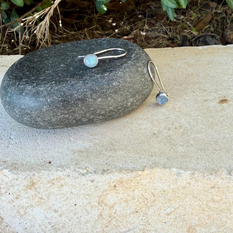 May include: A pair of silver earrings featuring round, iridescent moonstone cabochons set in metal bezels. The earrings have long, curved silver ear wires that extend from the back of the stone settings, creating a simple and elegant profile.