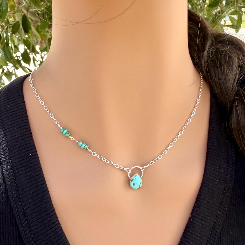 May include: A silver chain necklace featuring a small, teardrop-shaped turquoise pendant suspended from a circular silver ring. The chain is accented with three small, round turquoise beads on one side, creating a delicate and balanced design.