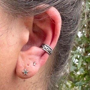 May include: Close-up of an ear adorned with silver jewelry. A braided ear cuff sits on the upper ear, while a star-shaped stud with a turquoise center and a small rectangular stud are in the earlobe. The background is blurred.
