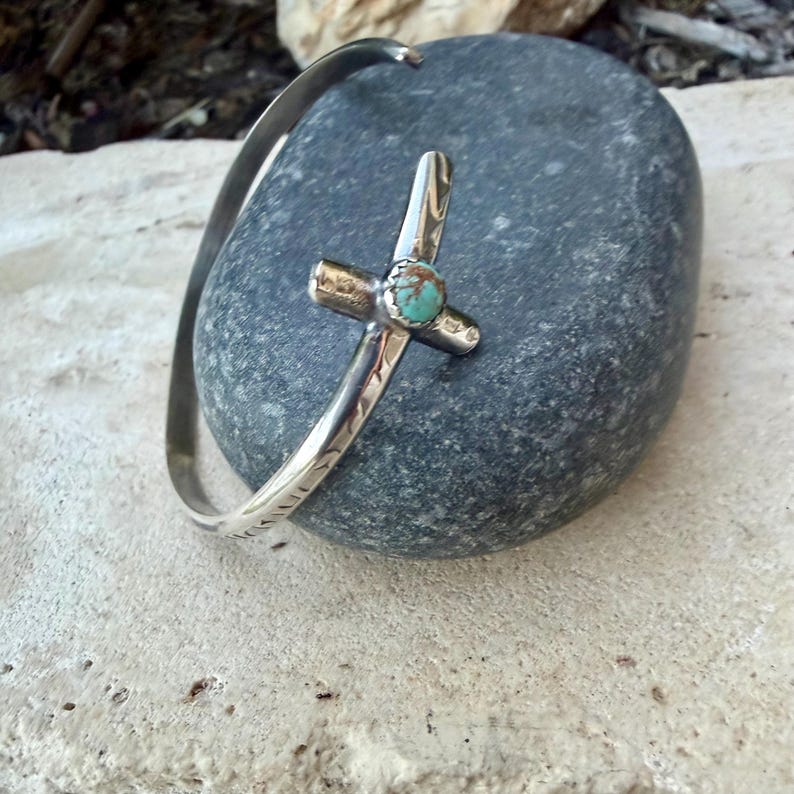 May include: A silver-toned cuff bracelet with a cross-shaped centerpiece. The cross features a small, round turquoise stone at its center. The bracelet is resting on a gray stone surface.