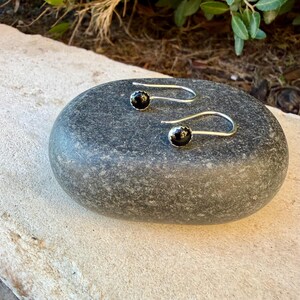 May include: A pair of silver hook earrings featuring small, round, polished black stones set in metal bezels. The earrings have a curved wire design that extends from the stone setting, creating a simple and clean look for these jewelry pieces.