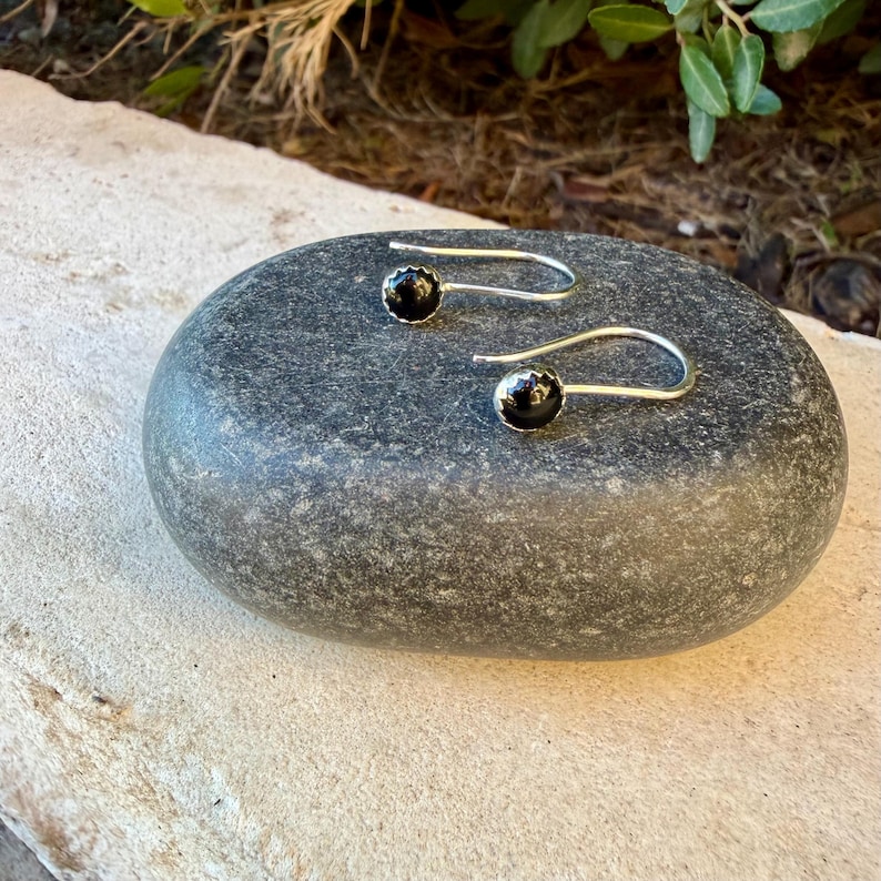 May include: A pair of silver hook earrings featuring small, round, polished black stones set in metal bezels. The earrings have a curved wire design that extends from the stone setting, creating a simple and clean look for these jewelry pieces.